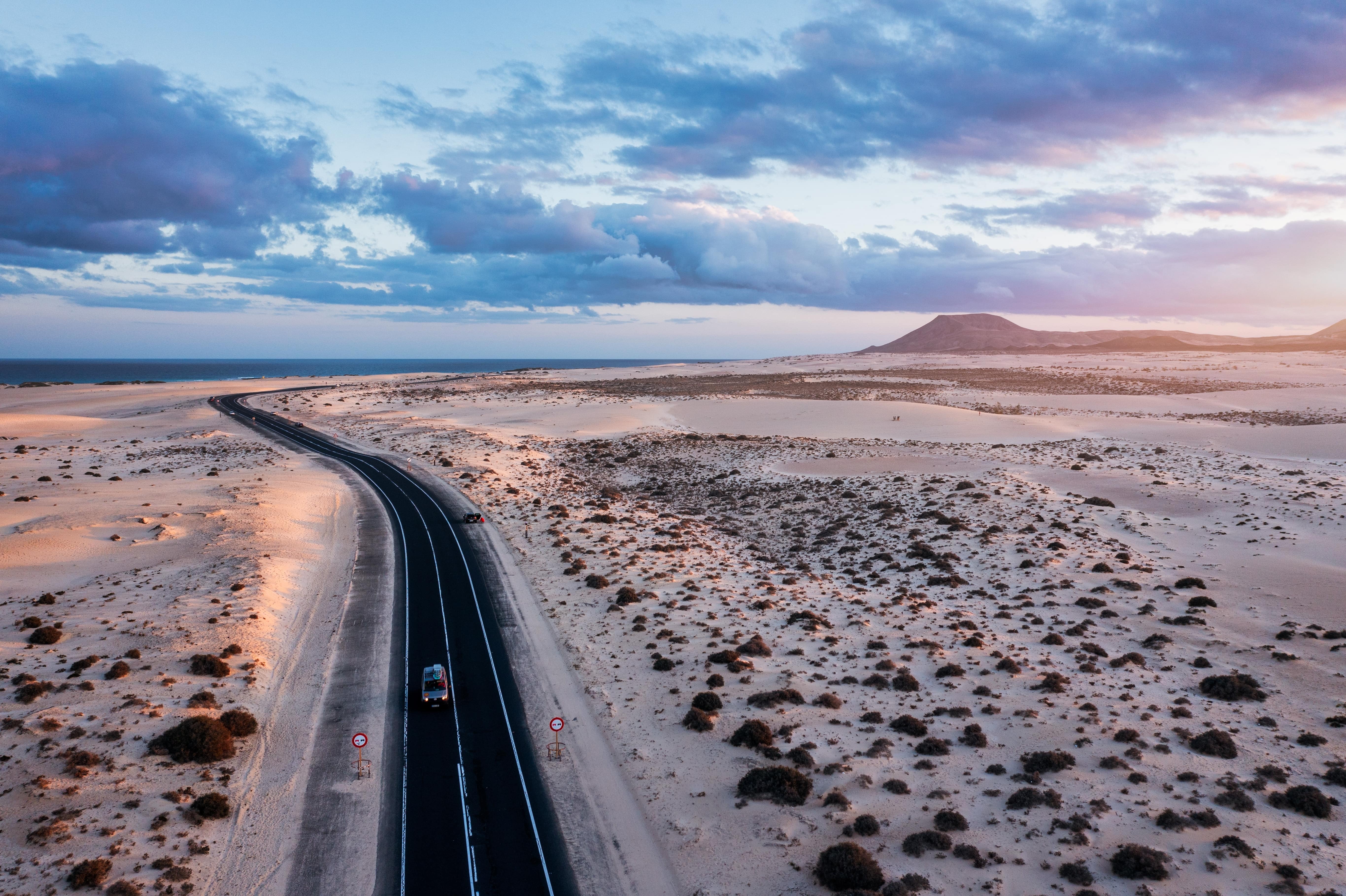 Route panoramique traversant les dunes de Corralejo à l'aube, avec l'océan en arrière-plan et un ciel teinté de rose et de bleu.